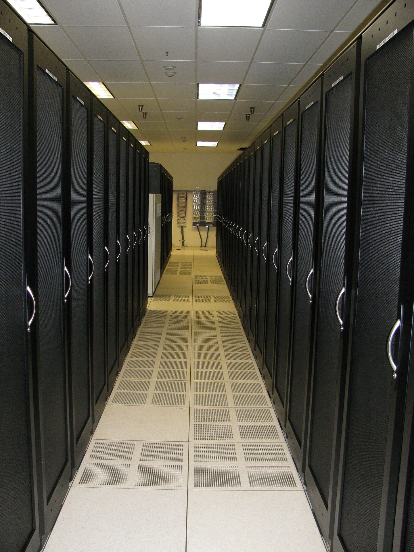 A datacenter aisle between two rows of black server cabinets with perforated mesh doors, standing on a raised floor with perforated floor tiles running down the middle of the aisle.