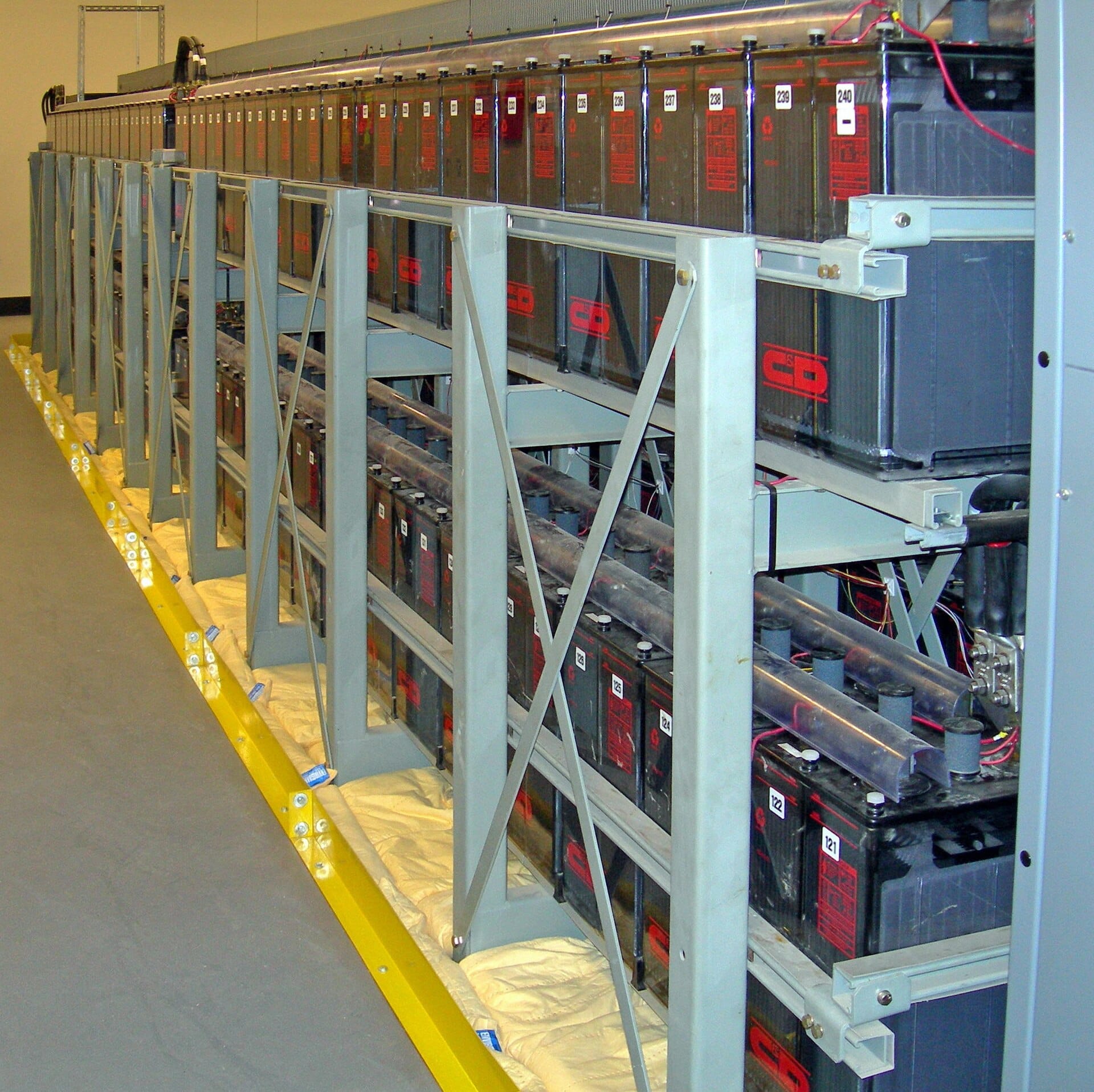 Rows of large sealed backup batteries mounted on metal racking inside a datacenter UPS room.