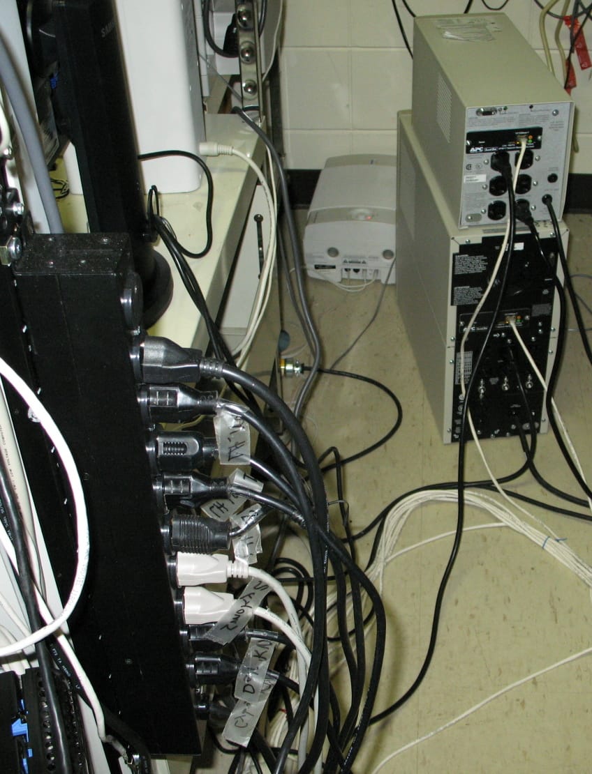 A cluttered equipment area with a black power strip covered in power plugs, each labelled with hand written masking tape, and unmanaged cables running across the floor to a UPS.