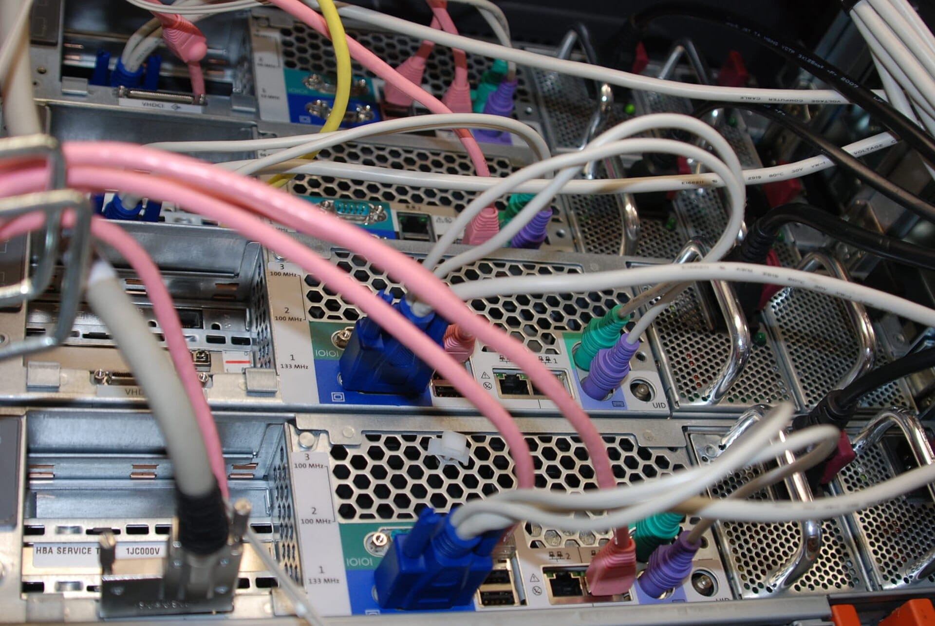 The rear of several rack mounted servers showing perforated power supply modules, network ports, a blue VGA connector, coloured cables plugged into each machine, and metal handles on the power supply units.
